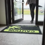 Detailne vaade mustale ohutusmatile, millel on erkkollane hoiatussümbol ja tekst "Caution Slippery When Wet", pakkudes selget visuaalset hoiatust märgade ja libedate põrandate korral