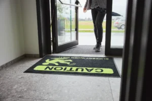 Detailne vaade mustale ohutusmatile, millel on erkkollane hoiatussümbol ja tekst "Caution Slippery When Wet", pakkudes selget visuaalset hoiatust märgade ja libedate põrandate korral