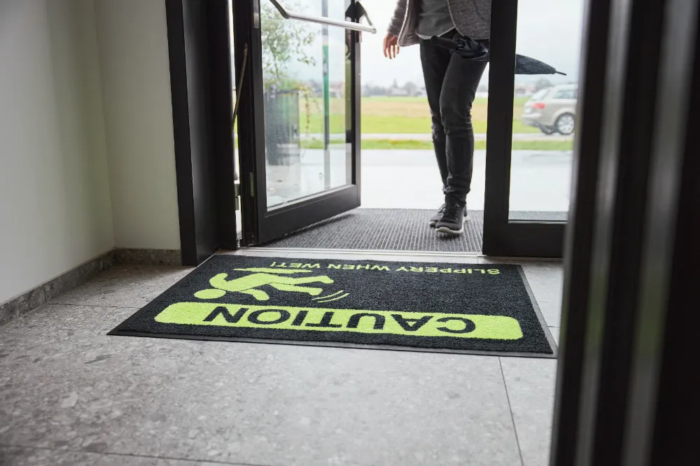 Detailne vaade mustale ohutusmatile, millel on erkkollane hoiatussümbol ja tekst "Caution Slippery When Wet", pakkudes selget visuaalset hoiatust märgade ja libedate põrandate korral