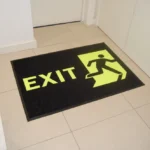 Musta värvi libisemiskindel ohutusmatt neoonrohelise Exit-märgistusega ja noolega, mis suunab külastajaid väljapääsu poole ja parandab siseruumides liikumisturvalisust