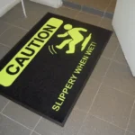 Must nailonist ohutusmatt, millele on trükitud neoonkollane hoiatussümbol ja tekst "Slippery when wet", et teavitada siseruumides liikujaid libisemisohtlikust põrandast