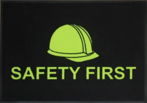 Musta värvi nailonist ohutusmatt erkkollase "Safety First" märgisega, mis on loodud tööstusruumidesse ja ladudesse, et rõhutada tööohutust ja vältida libastumisi