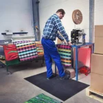 Töötaja seismas Floorstudio ergonoomilisel kummimatil pakkimislaua taga väsimuse leevendamiseks