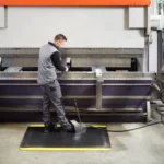 Ergonoomilised matid 43 Tööline seadistamas suurt tööstuslikku pinki, seistes kollaste servadega ergonoomilisel matil