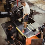 Ergonoomilised matid 54 Tööline seismas platvormil kollaste servadega FloorStudio ergonoomilisel matil rasketööstuses, sädemed lendamas