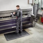 Ergonoomilised matid 55 Hallis tööriietuses mees seismas mustal FloorStudio ergonoomilisel matil suure metallipainutuspingi ees