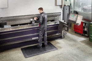 Hallis tööriietuses mees seismas mustal FloorStudio ergonoomilisel matil suure metallipainutuspingi ees