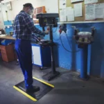 Ergonoomilised matid 57 Kaitsevarustuses töömees töötamas puurpingi taga, seistes FloorStudio kollaste ohutusservadega ergonoomilisel matil