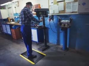Kaitsevarustuses töömees töötamas puurpingi taga, seistes FloorStudio kollaste ohutusservadega ergonoomilisel matil