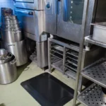 Ergonoomilised matid 58 Must FloorStudio ergonoomiline matt paigaldatuna suurköögi roostevabast terasest ahjude ja riiulite ette