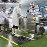 Ergonoomilised matid 67 Kolm toiduainetööstuse töötajat valgetes kitlites ja kummikutes seismas kollaste servadega FloorStudio kummimattidel töölaua taga