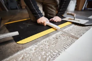 Must ristkülikukujuline FloorStudio ergonoomiline matt (85x150 cm) betoonpõrandal töömehe käte all