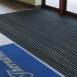 Harjasmatid 13 Alumiiniumist porirest ja sinised logomatid ärihoone klaasukse ees sissekäigul