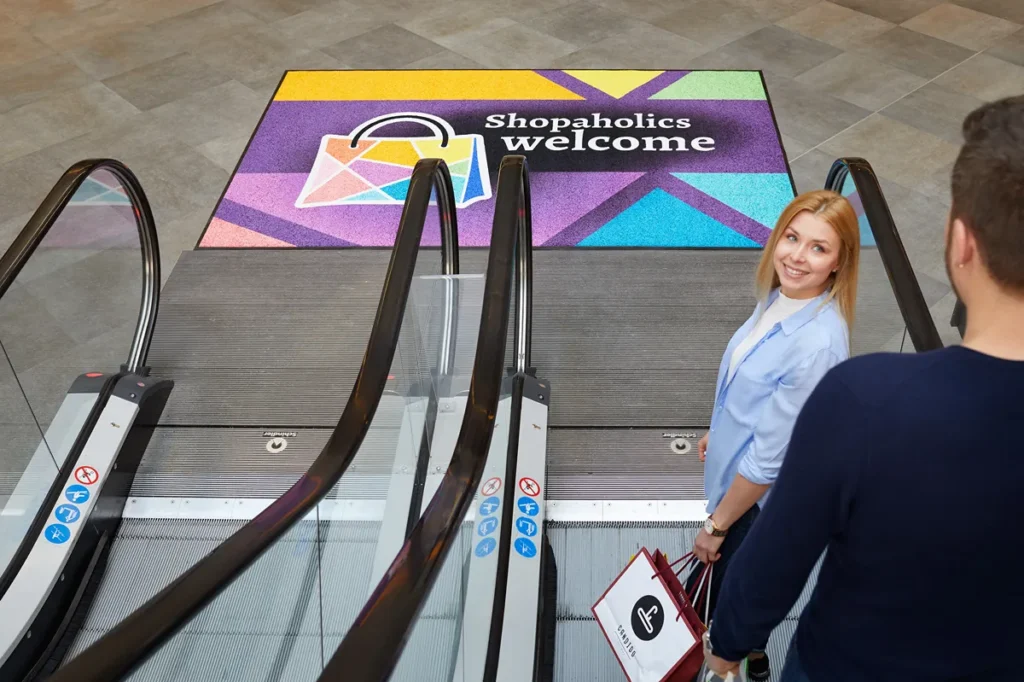 Kohandatud disainiga logovaip tekstiga "Shopaholics welcome" kaubanduskeskuse sissepääsus eskalaatori ees