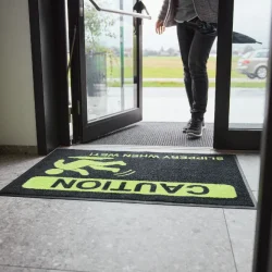 Detailne vaade mustale ohutusmatile, millel on erkkollane hoiatussümbol ja tekst "Caution Slippery When Wet", pakkudes selget visuaalset hoiatust märgade ja libedate põrandate korral